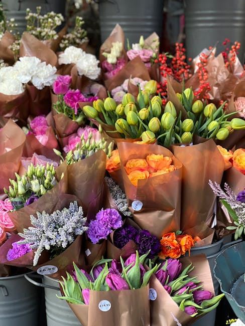 A vibrant display of freshly cut flowers arranged in brown paper wrapping, including white carnations, pink and purple blooms, green-tipped tulips, orange roses, and purple statice. The bouquets are n