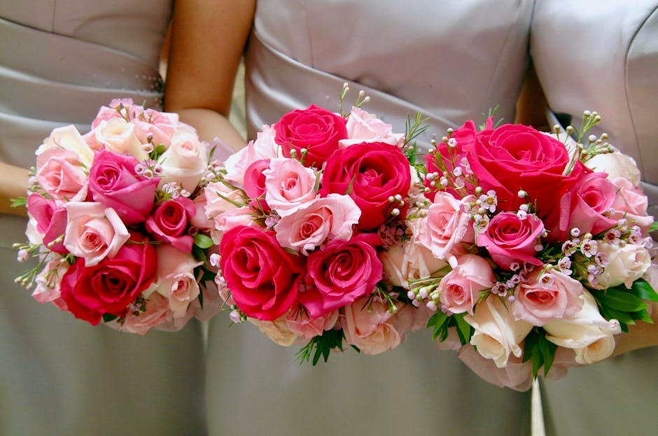 A floral arrangement featuring three bouquets of fresh roses in shades of pink, pale pink, and deep red, wrapped with white and pale pink tissue paper and tied with soft pink ribbons. The roses are ar