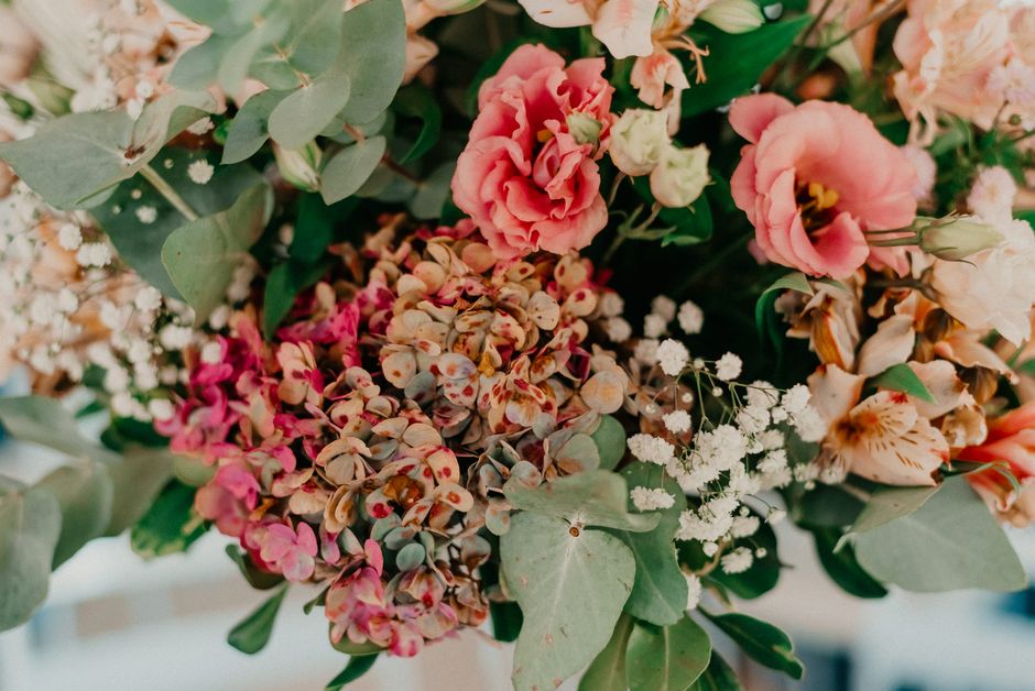 A close-up of a floral arrangement featuring soft pink and peach roses, white baby's breath, and dusty miller foliage. The arrangement is holding a mix of small, delicate flowers and broad, muted gree