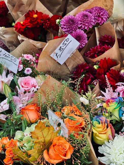 A vibrant assortment of fresh flowers including red dahlias, purple chrysanthemums, pink roses, orange roses, and multicolored rainbow roses arranged in brown paper wrapping. The bouquets are lush and
