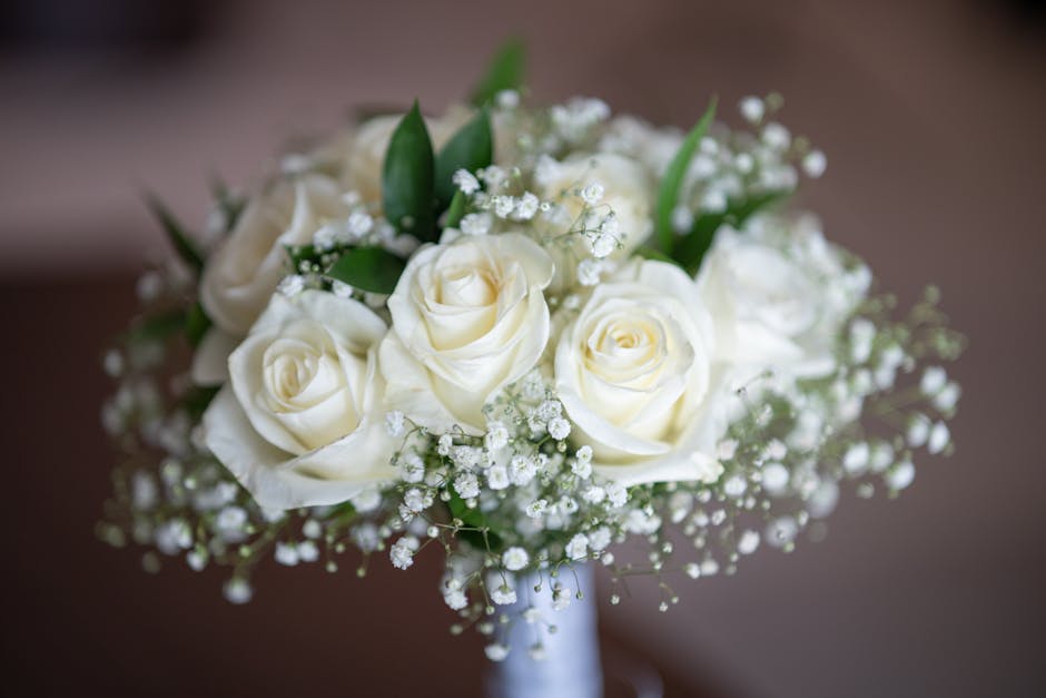 A close-up image of a bridal bouquet featuring ivory roses with softly curved petals, complemented by delicate white baby's breath scattered throughout and accented with glossy green leaves. The bouqu