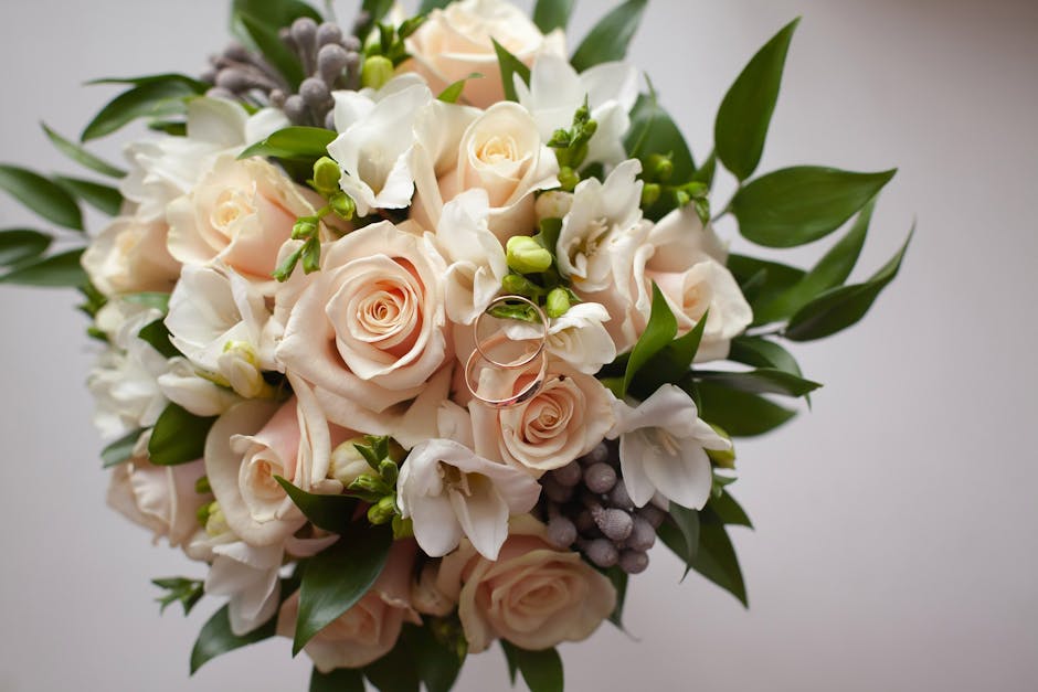 A fresh floral bouquet featuring light blush pink roses, white freesias, and small green buds, accented with lush green leaves and some clusters of greyish lavender berries. The flowers are arranged i