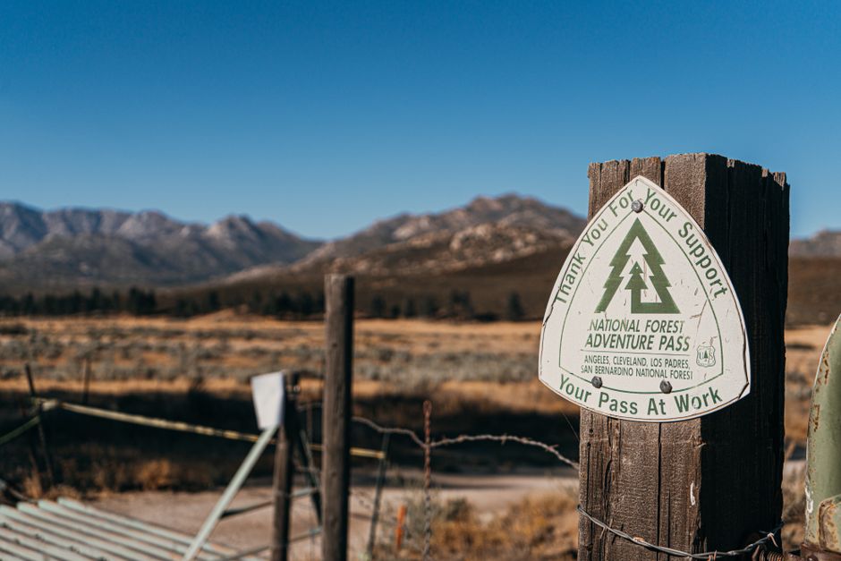A close-up of a wooden post displaying a National Forest Adventure Pass sticker with green forestry symbols and text, set against a scenic background of distant mountains, sparse vegetation, and a cle