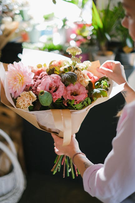 A person holding a large floral bouquet featuring soft pink peonies, cream carnations, and delicate white flowers, accented with dark green foliage. The bouquet is wrapped in cream and white paper wit